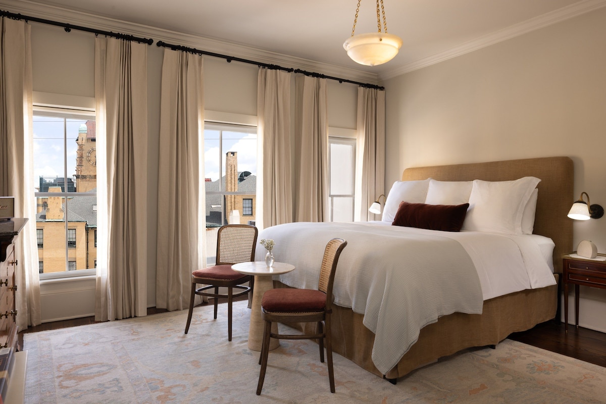 Habitación de hotel con una cama grande, una mesita, una silla y vistas a la ciudad a través de las ventanas.