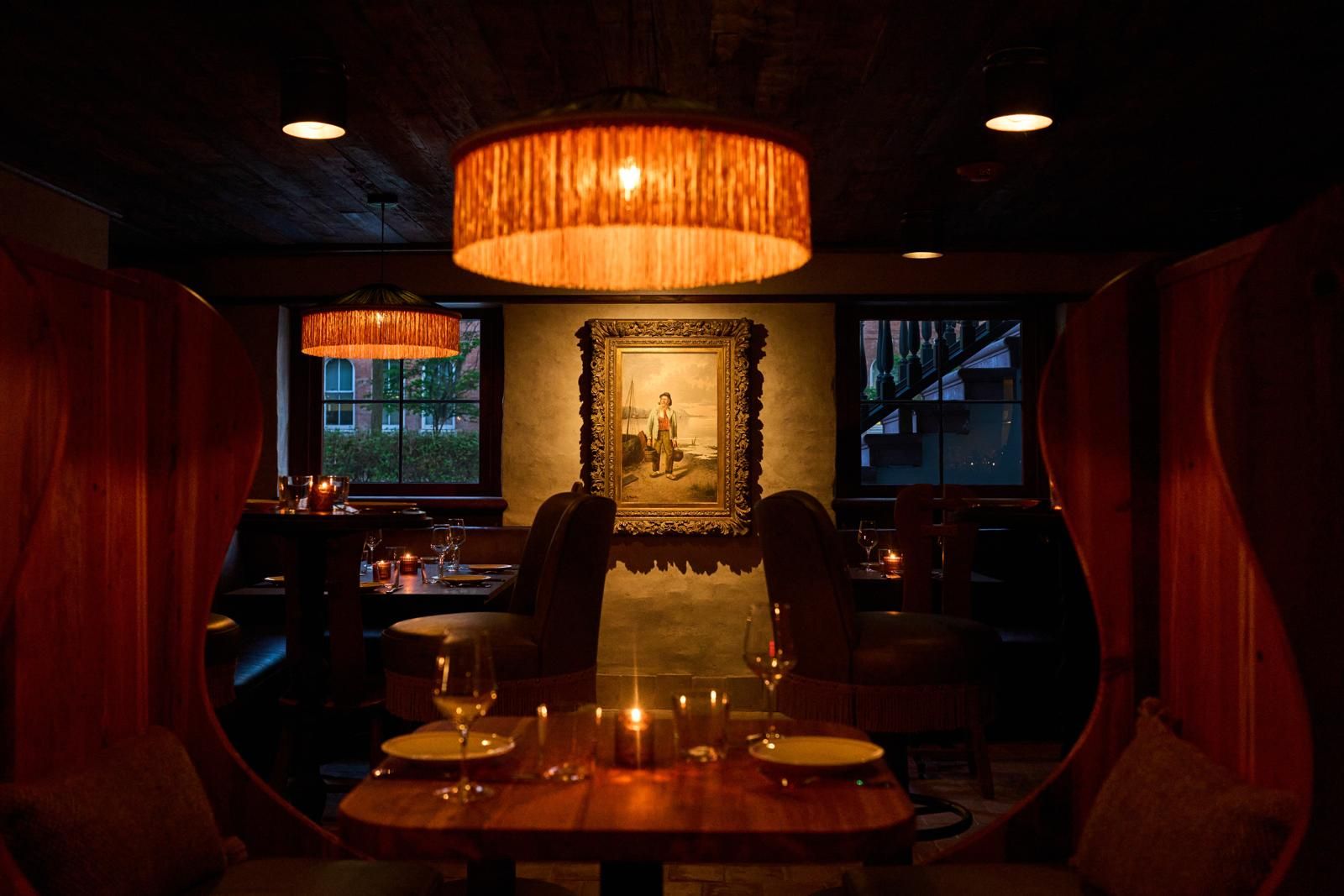 An interior with a dining table set for two, surrounded by chairs, under a large hanging lamp.