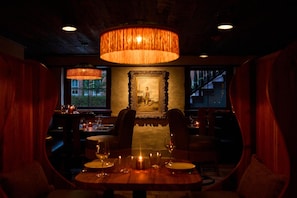 An interior with a dining table set for two, surrounded by chairs, under a large hanging lamp.