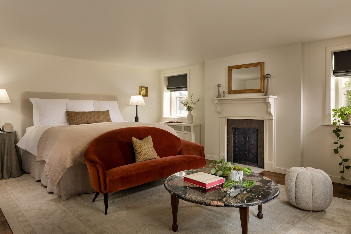 Un dormitorio con una cama, un sillón rojo, una mesita redonda con un libro, una chimenea y una ventana con una planta.