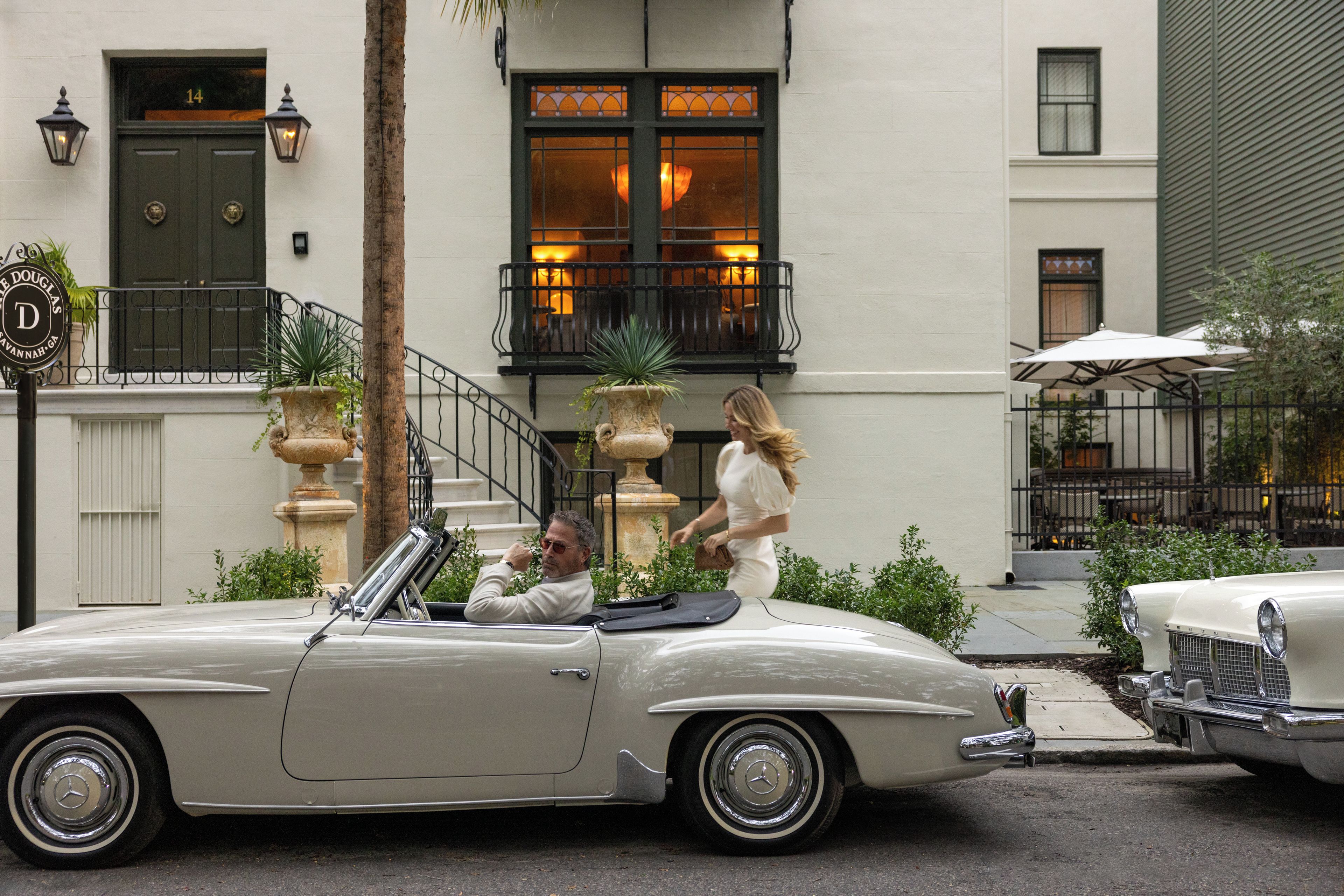 A classic convertible car parked in front of a building with a stone facade, a staircase, and a balcony with a railing.