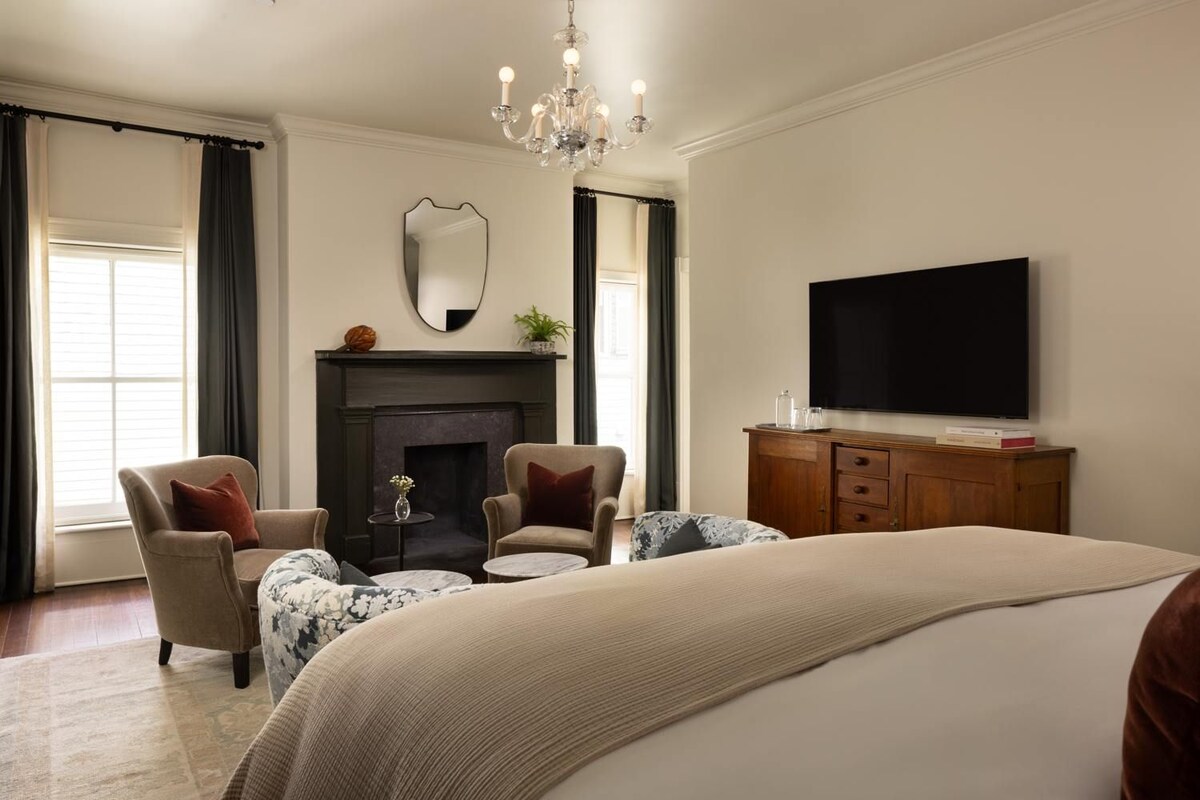 Une chambre d’hôtel avec un grand lit, deux fauteuils, une cheminée, une télévision et un lustre.