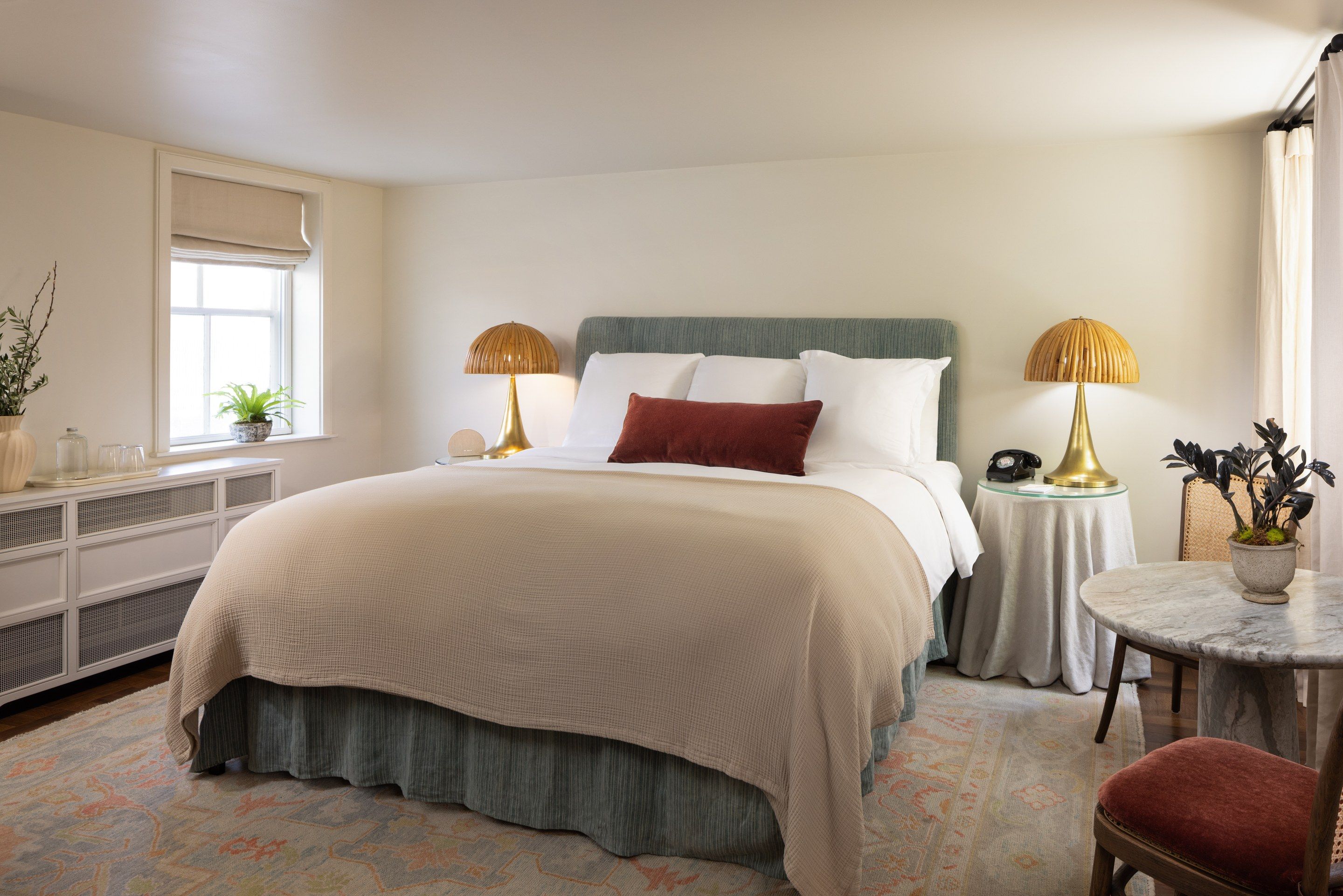 A bedroom with a bed, bedside tables, a nightstand, a chair, a small table, a potted plant, and a window.