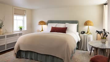 A bedroom with a bed, bedside tables, a nightstand, a chair, a small table, a potted plant, and a window.