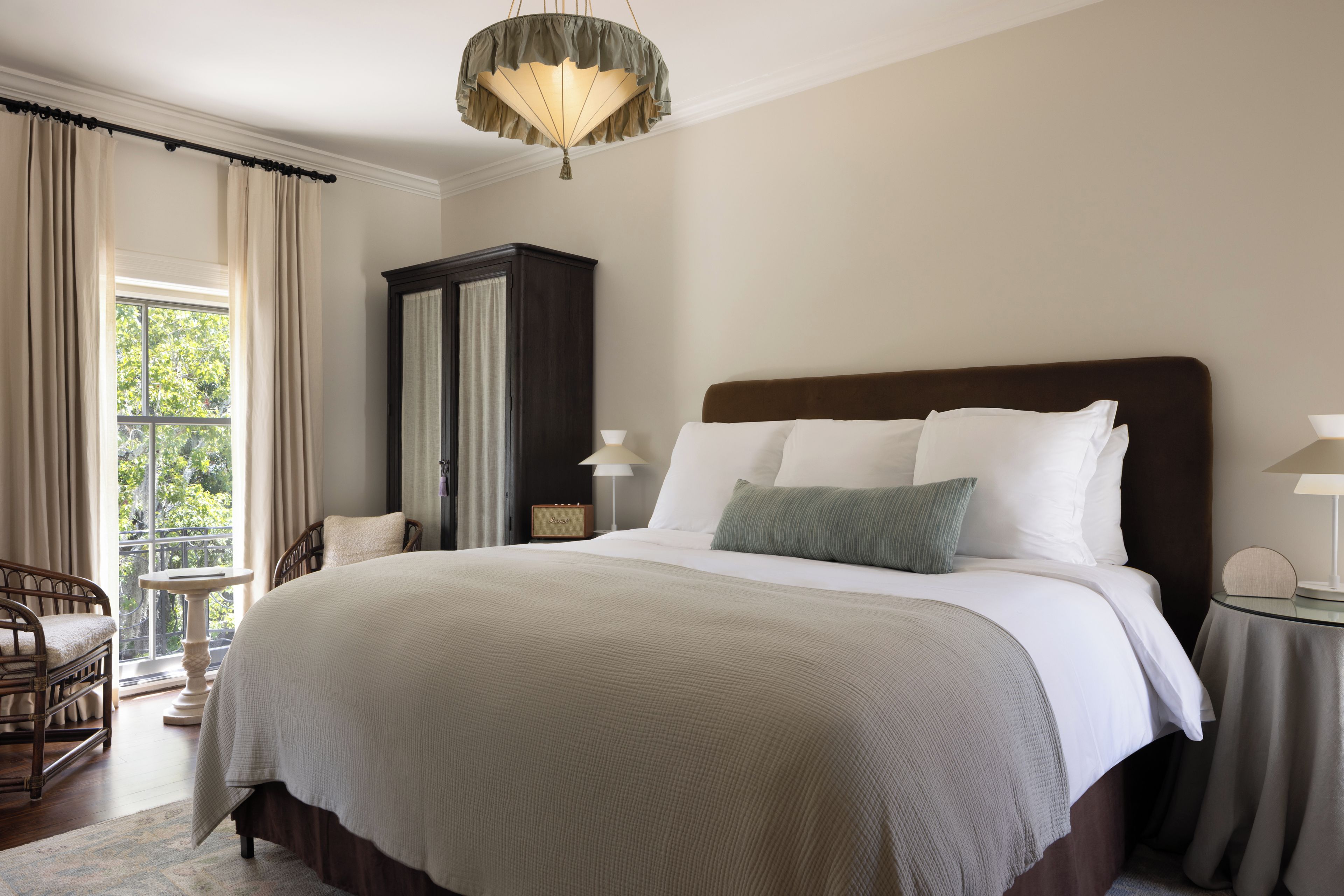 A hotel room with a large bed, a wooden wardrobe, a sitting area with a chair, and a view through a window.