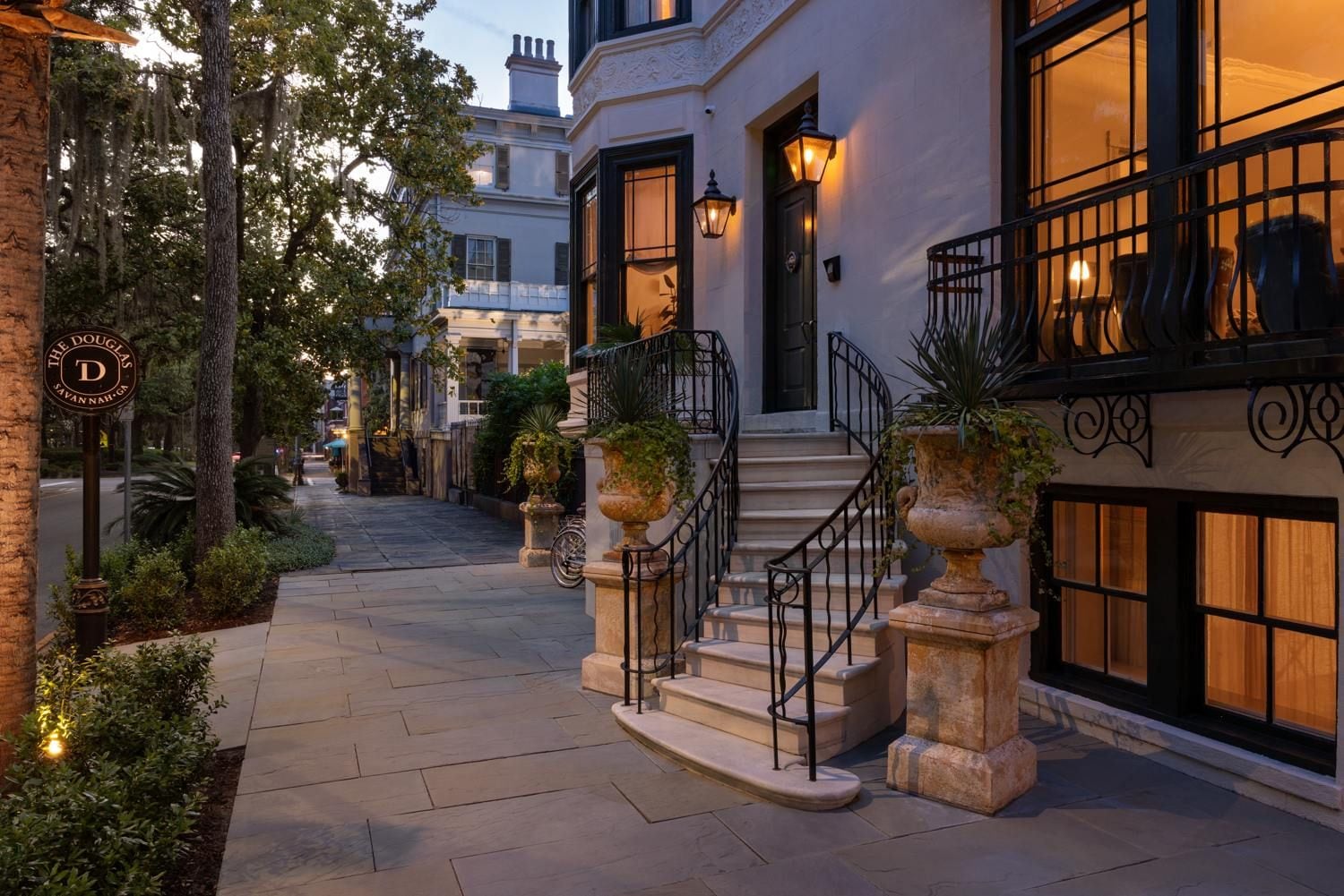 A well-lit hotel entrance with a stone staircase leading to the door, surrounded by potted plants and a paved walkway.