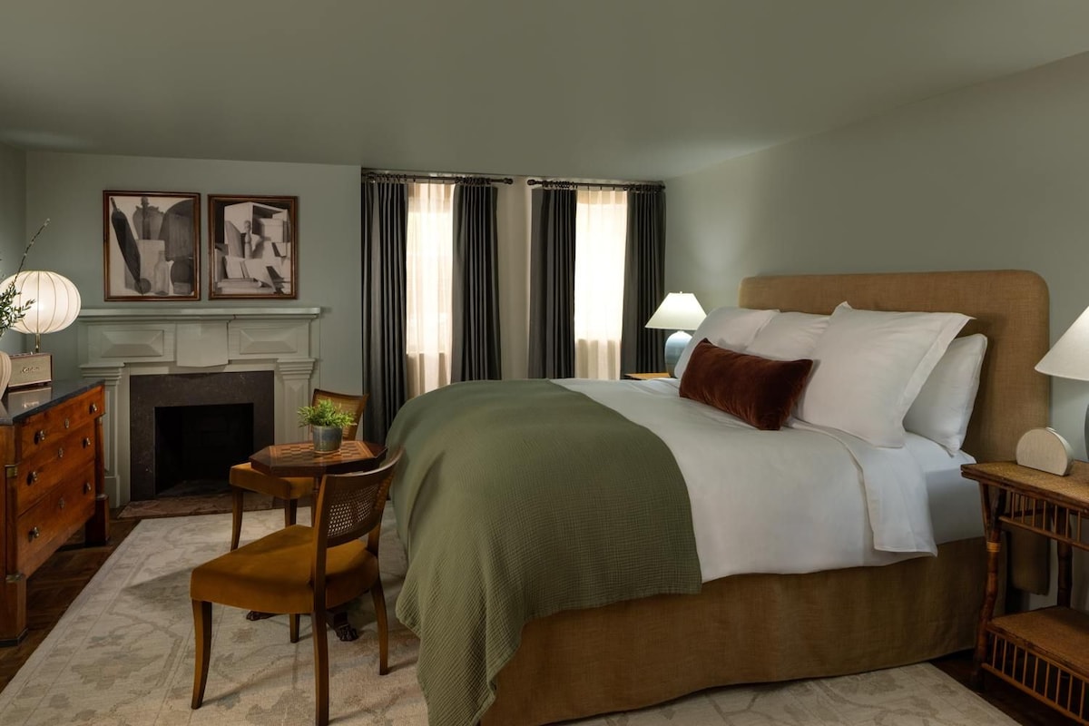 Une chambre d’hôtel avec un lit, une cheminée, une table à manger avec des chaises et des œuvres d’art accrochées aux murs.
