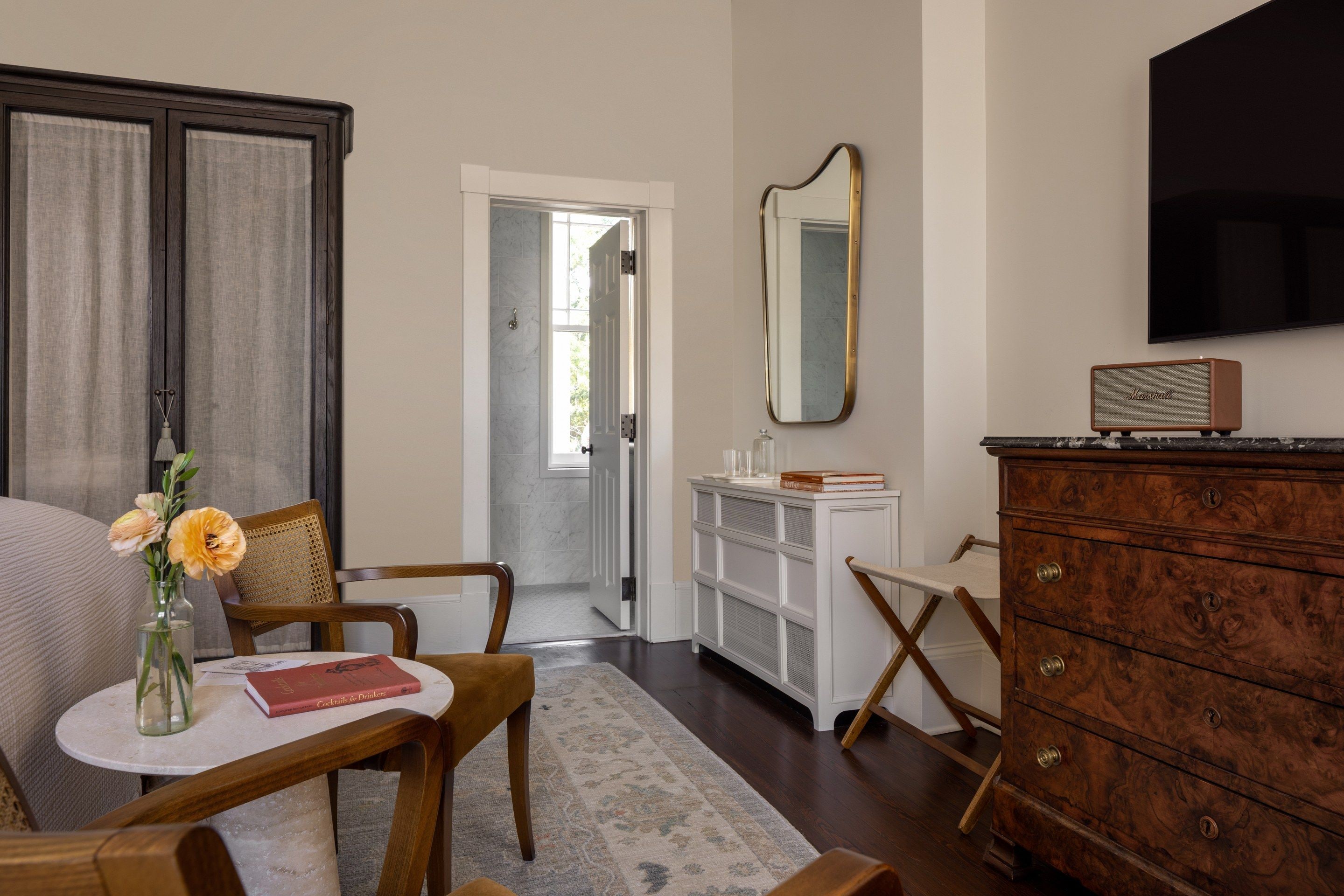 A room with a bed, a table with a book and flowers, a dresser, a mirror, and a television.