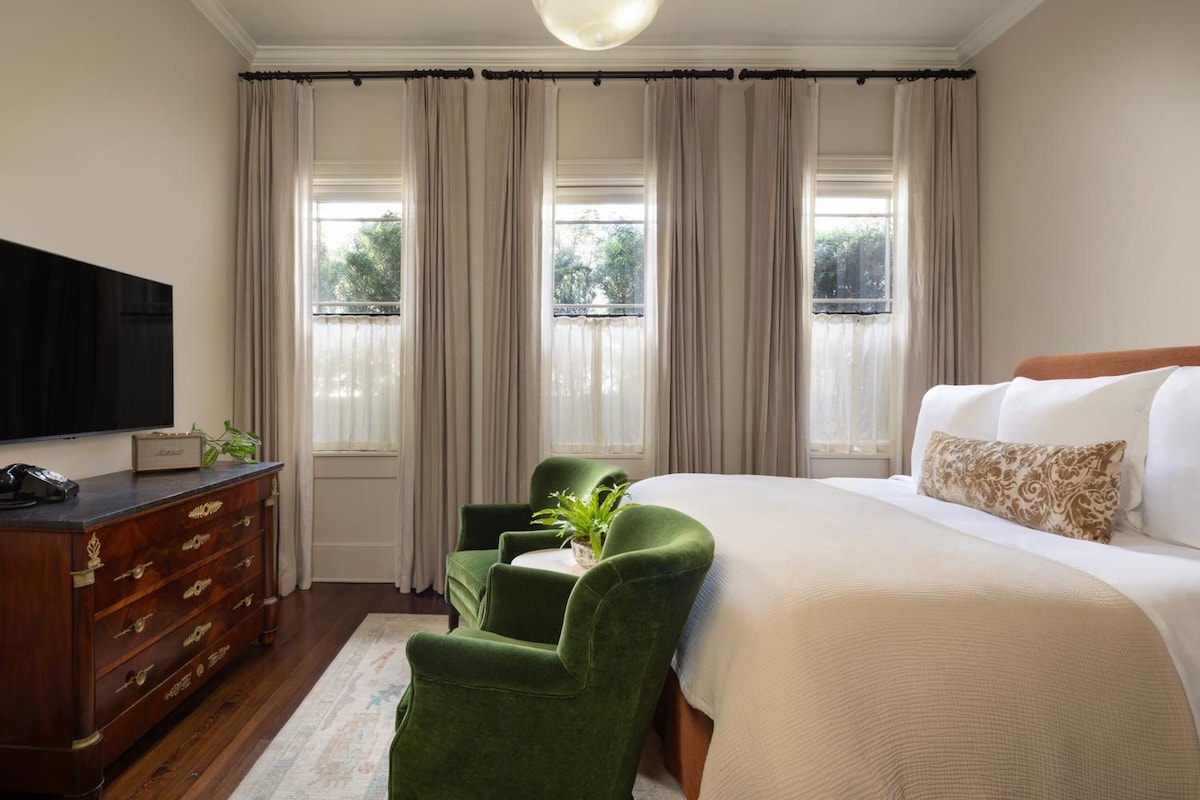 Une chambre d’hôtel avec un grand lit, un téléviseur posé sur une commode en bois et deux fauteuils verts.