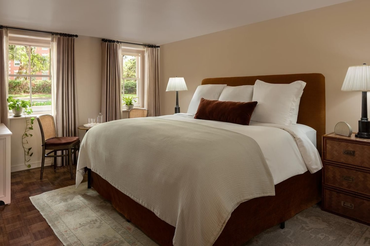 Une chambre d’hôtel avec un grand lit, des lampes de chevet, une chaise, une petite table et une fenêtre avec des rideaux.