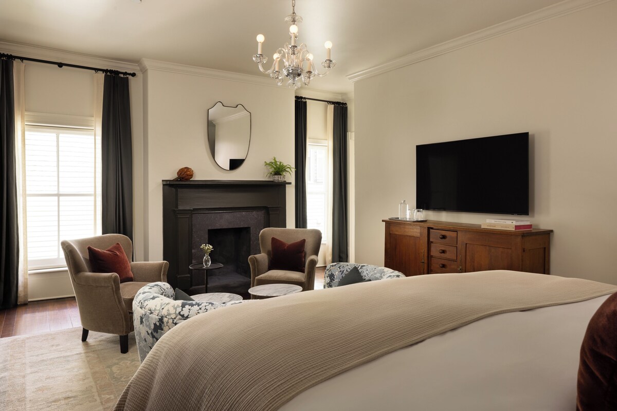 A bedroom with a large bed, two armchairs, a fireplace, a television, and a chandelier.