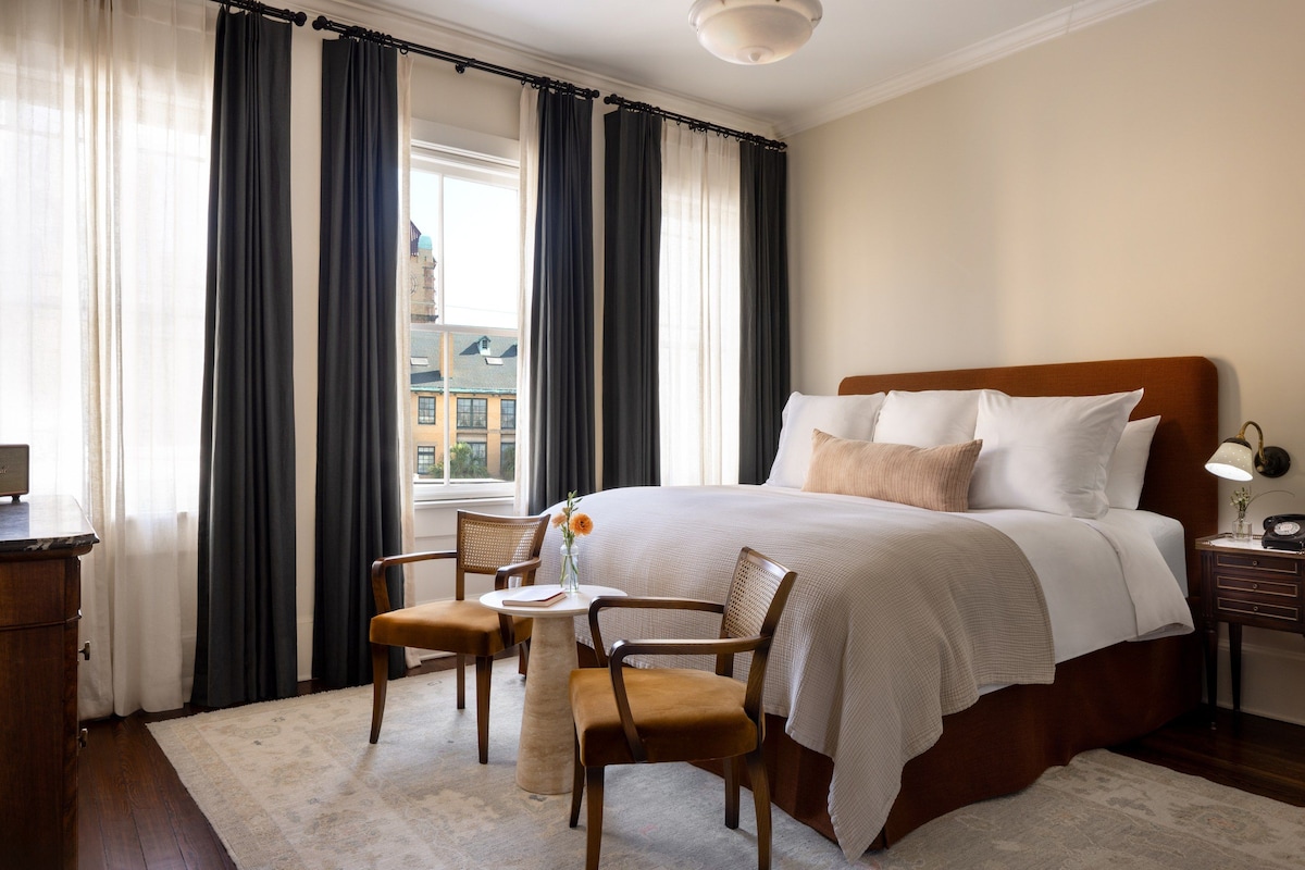 Habitación de hotel con una cama grande, dos sillas, una mesita y una mesita de noche con una lámpara.