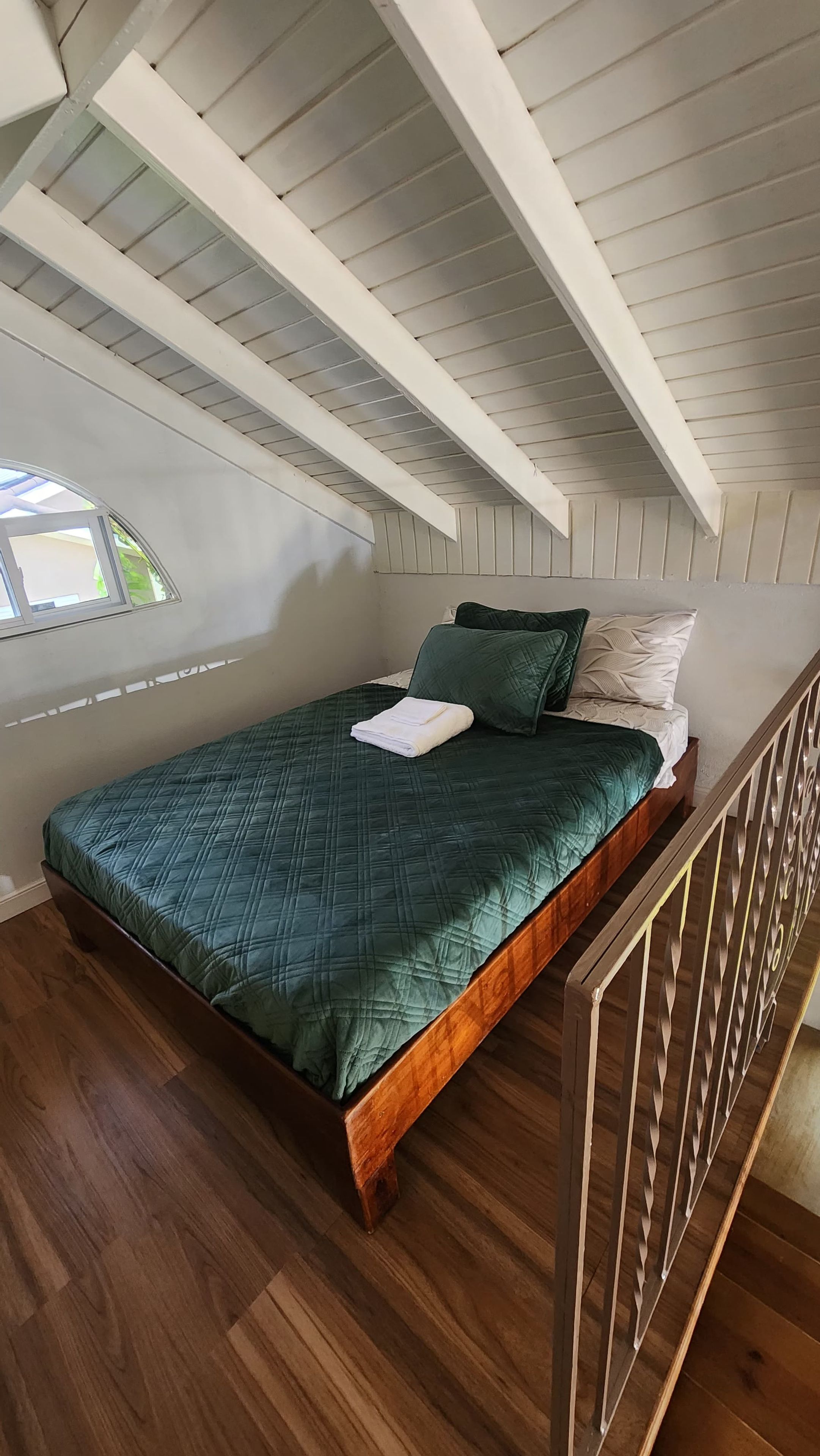 Un lit simple avec une couette verte, un oreiller blanc et un oreiller vert foncé, dans une pièce au plancher en bois et au plafond en pente.