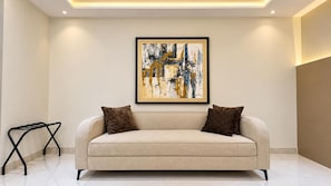 A modern living room with a beige sofa, two dark cushions, a black metal side table, and a framed abstract painting on the wall. - The Barli Hotel and Apartments (Abuja)