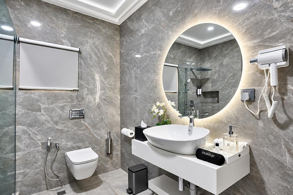 A modern bathroom with a large oval sink, a round mirror, a handheld showerhead, and a hairdryer mounted on the wall.