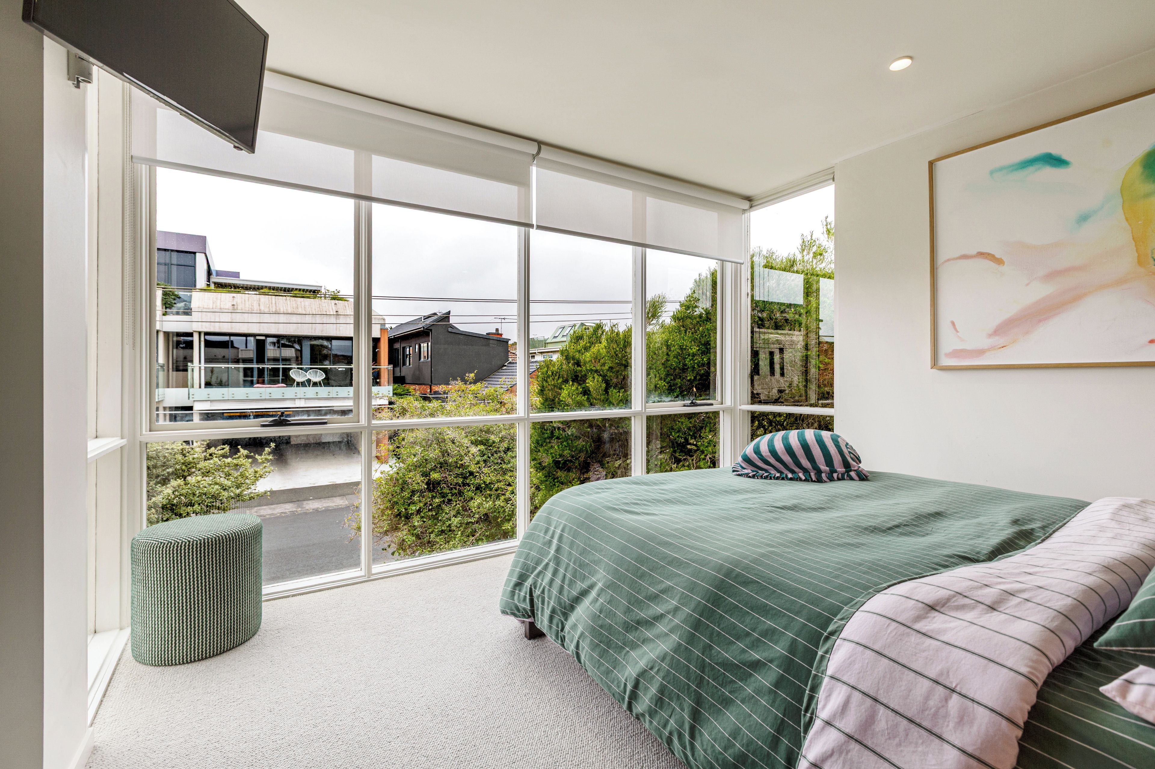 Ein Schlafzimmer mit großem Fenster, einem Bett mit grüner Bettwäsche, einem kleinen Hocker und einem Gemälde an der Wand.