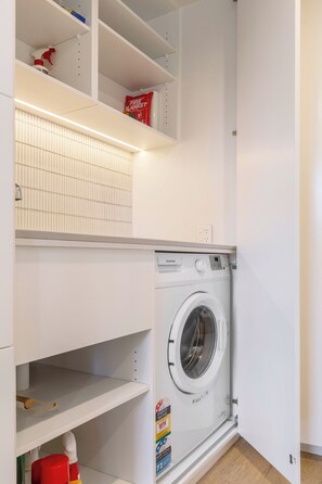 A laundry room with a front-loading washing machine, shelves above and below, and a fire blanket on the top shelf. - Bayside Grove The Heart of St Kildas Coastal Life (Saint Kilda)