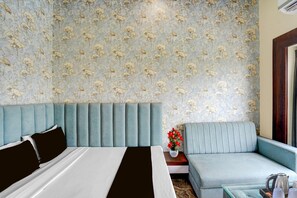 A hotel room with a bed, a sofa, a nightstand with a vase of flowers, and a wall with floral wallpaper. - Collection O Narsi Vihar Park Deluxe (Mathura)