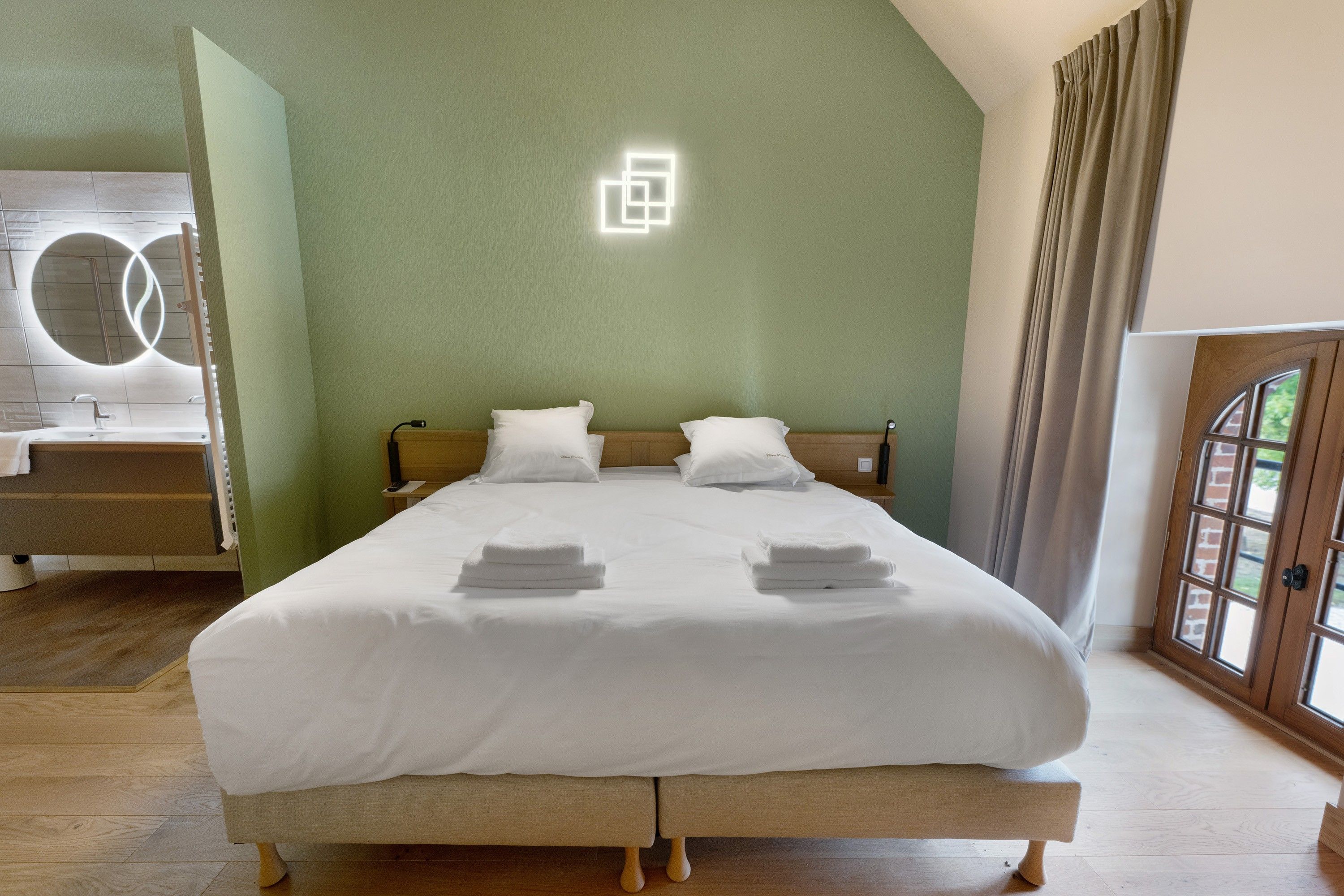 Habitació d'hotel amb un llit, un panell de capçalera de fusta, una tauleta de nit, un mirall i una finestra amb cortines.