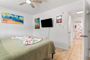 Une pièce comprenant un lit, un ventilateur de plafond, un téléviseur et une porte donnant sur une autre pièce.