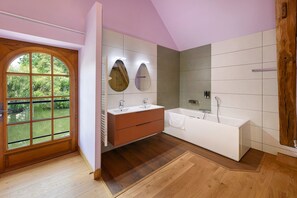 Ein modernes Badezimmer mit Holztür, großem Fenster, Waschtisch mit zwei Becken, Badewanne und einer Wandvertäfelung aus Holz.