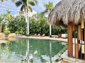 Ein Poolbereich mit Liegestühlen, einer Struktur mit Reetdach und tropischer Vegetation.