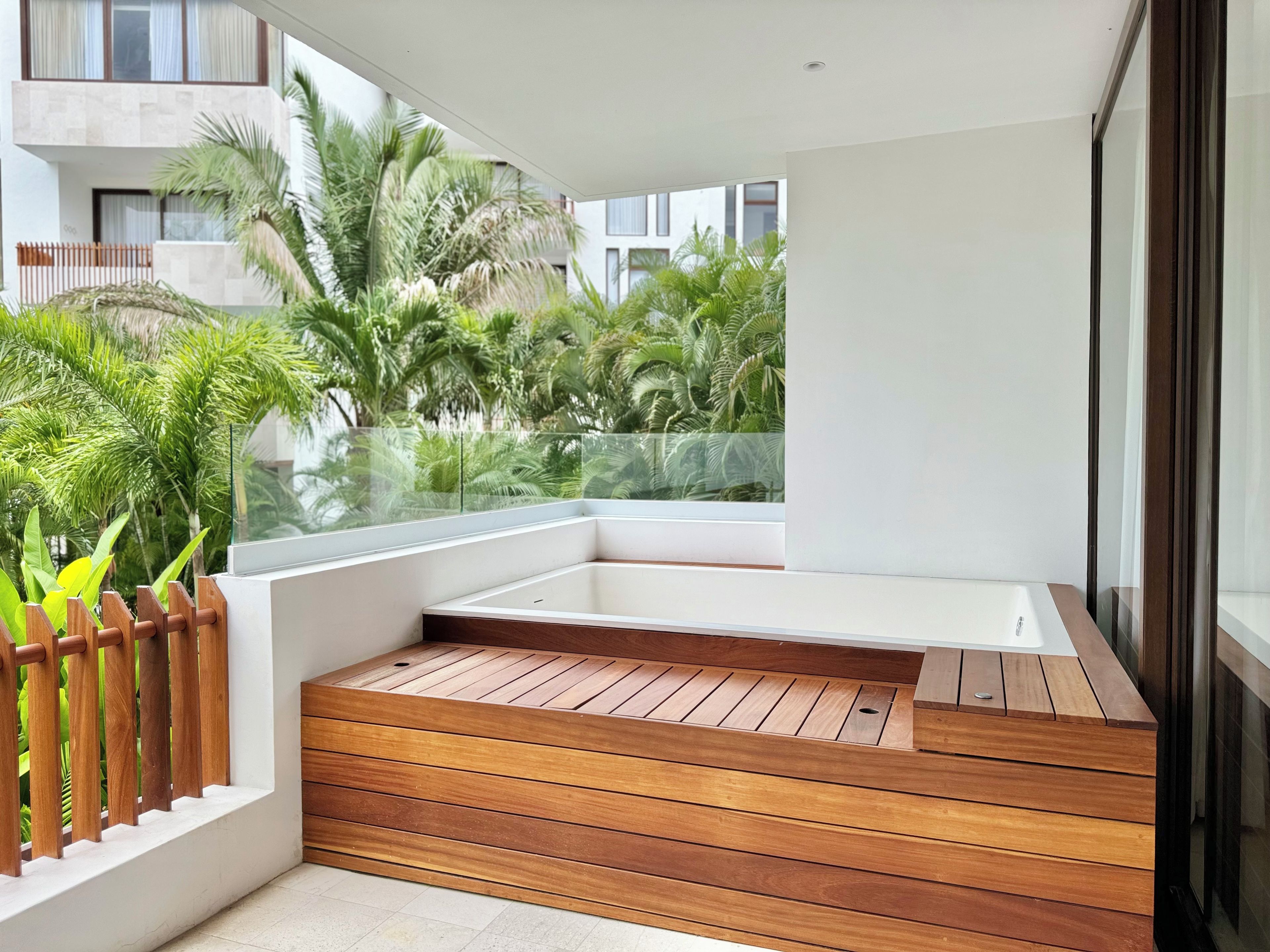 Un balcone moderno con pavimento in legno, ringhiera in vetro e vista su vegetazione ed edifici.
