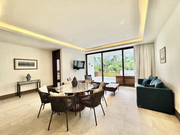 Ein modernes Wohnzimmer mit Essbereich, einer Couch, einem an der Wand montierten Fernseher und einem Balkon mit Blick auf Grünflächen.