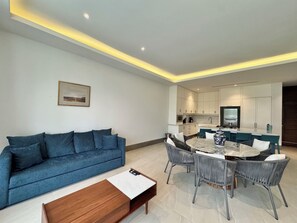 Een moderne woonkamer met een blauwe bank, een houten salontafel en een eethoek met een marmeren tafel en rieten stoelen.