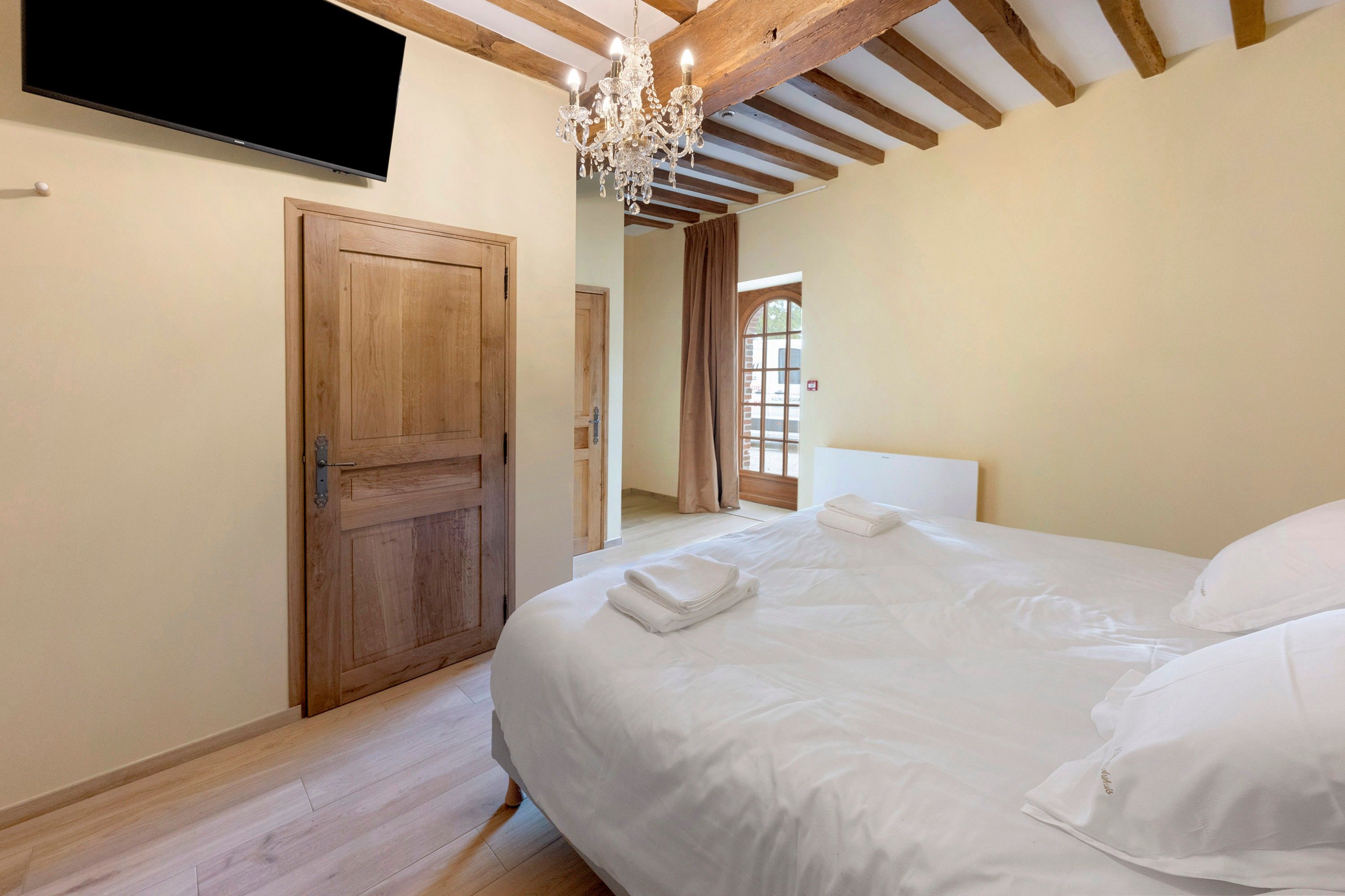 Ein Schlafzimmer mit Holztür, einem an der Wand montierten Flachbildfernseher, einer Kristallleuchtre und einem Bett mit weißer Bettwäsche.