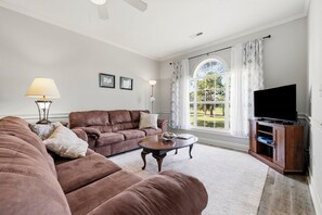 Ein Wohnzimmer mit einem braunen Sofa, einem Couchtisch, einer Lampe, einem Fernseher und einem großen Fenster mit Vorhängen.