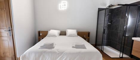 Une chambre d’hôtel avec un lit, une porte en bois, un espace douche et un luminaire fixé au mur.