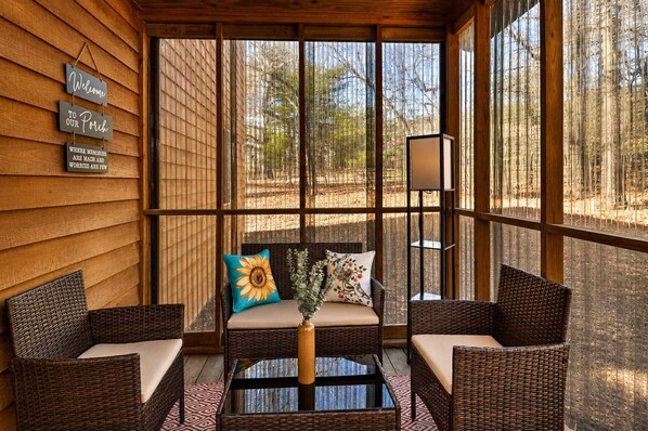 Eine Veranda mit Korbmöbeln, einem Glastisch und Blick auf Bäume durch große Fenster.