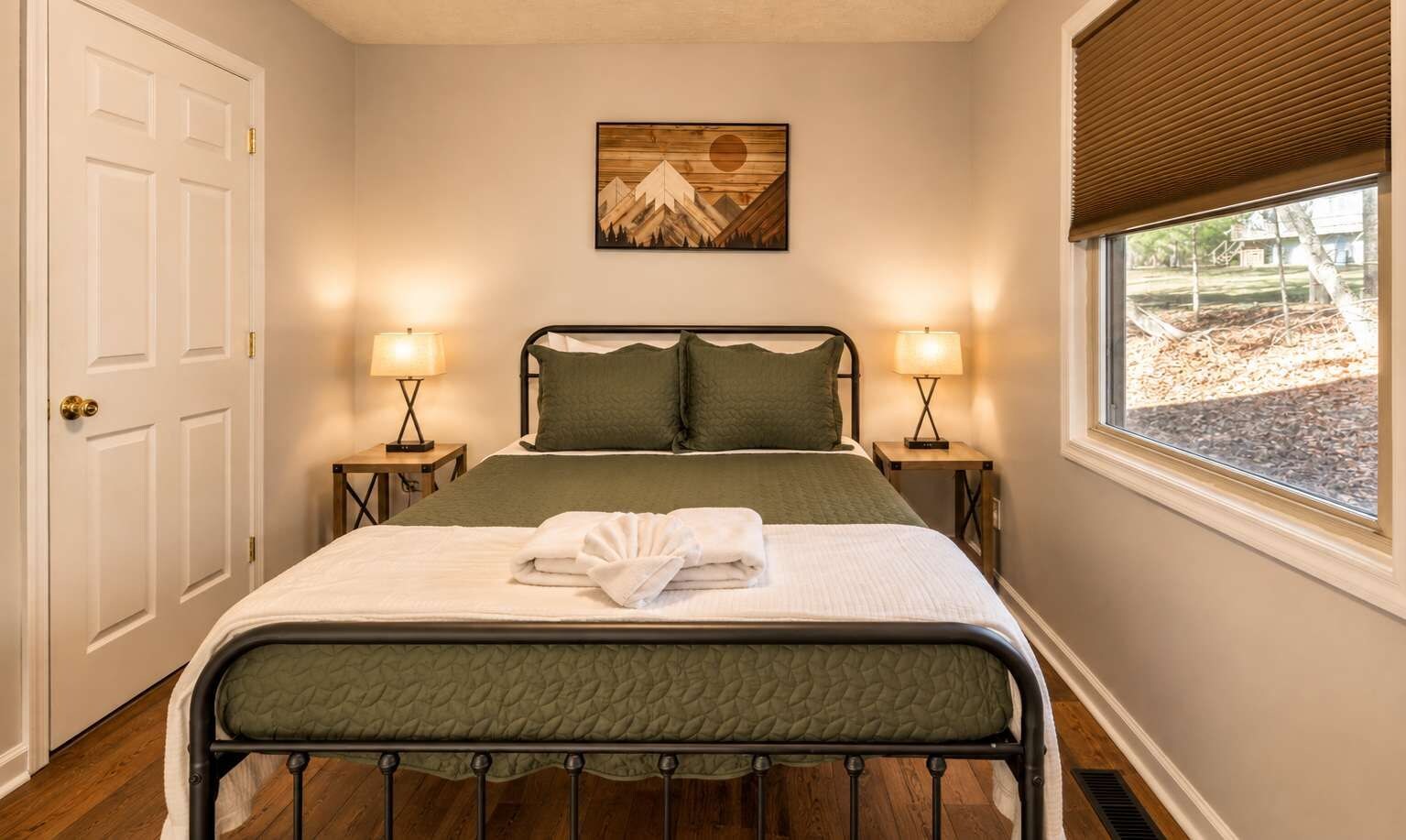 A neatly made bed with a green bedspread and pillows, two bedside lamps, a door, and a window with blinds.
