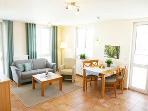 A living room with a sofa, coffee table, and dining table with chairs. There is a radiator, a lamp, and a window with curtains. - Apartment Building Linquenda 14 (Heringsdorf)