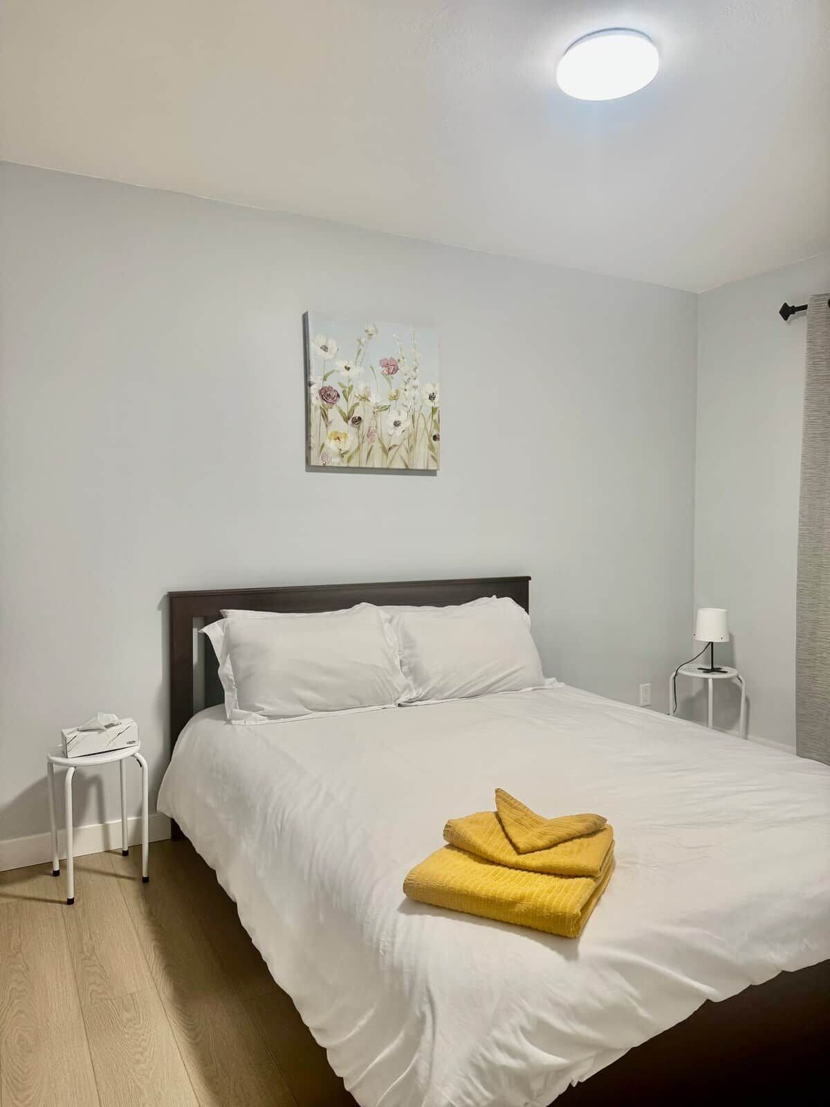 Une chambre à coucher avec un lit, du linge de lit blanc, une table de chevet avec une lampe et un tableau au mur.