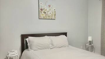 Une chambre à coucher avec un lit, du linge de lit blanc, une table de chevet avec une lampe et un tableau au mur.