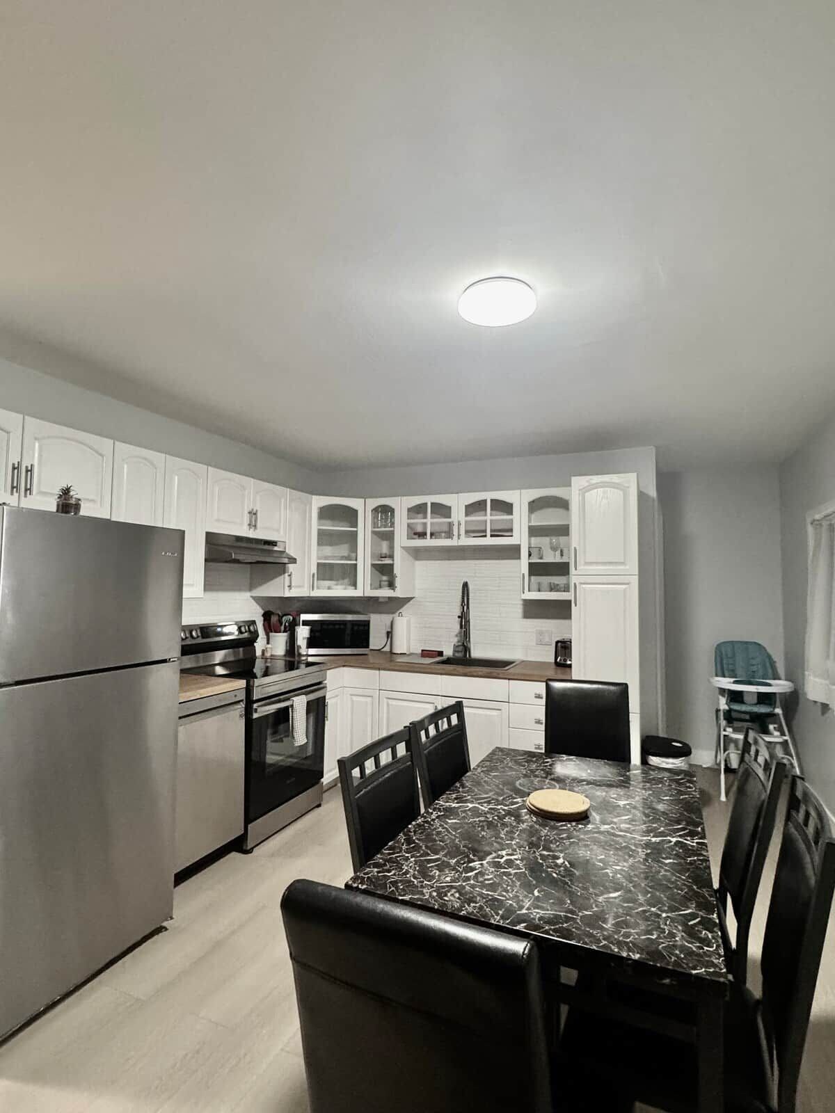 A modern kitchen with white cabinets, stainless steel appliances, a marble countertop, and a dining area with black chairs.
