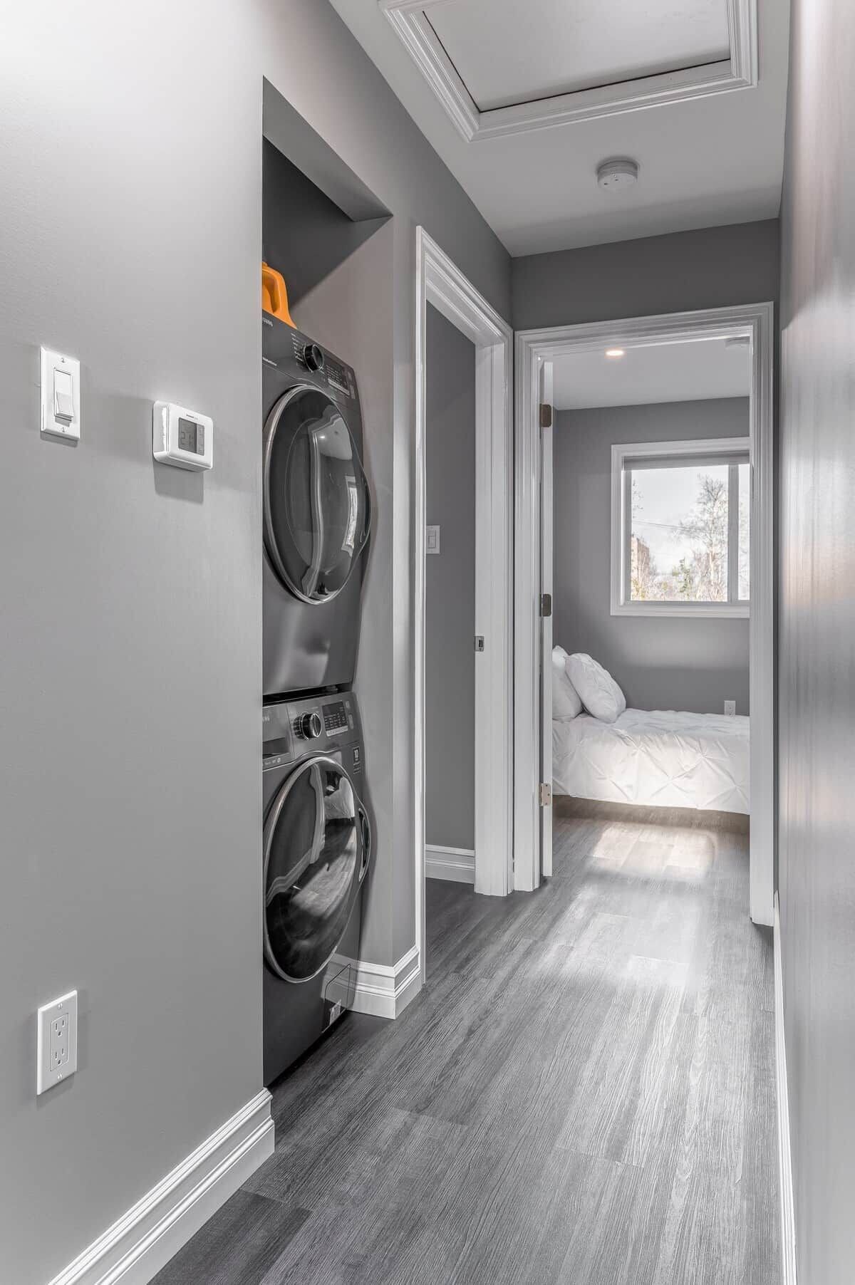 A laundry room with stacked washers and dryers, a bed in the adjacent bedroom, and a window providing natural light.