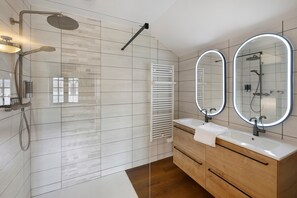 Ein modernes Badezimmer mit einer Duschkabine aus Glas, einem weißen Handtuch am Waschbecken und zwei beleuchteten, an der Wand befestigten Spiegeln.