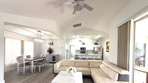 Ein modernes Wohnzimmer mit einer Eckcouch, einem Essbereich mit Tisch und Stühlen, einer Küche mit weißen Schränken und einem Deckenventilator.