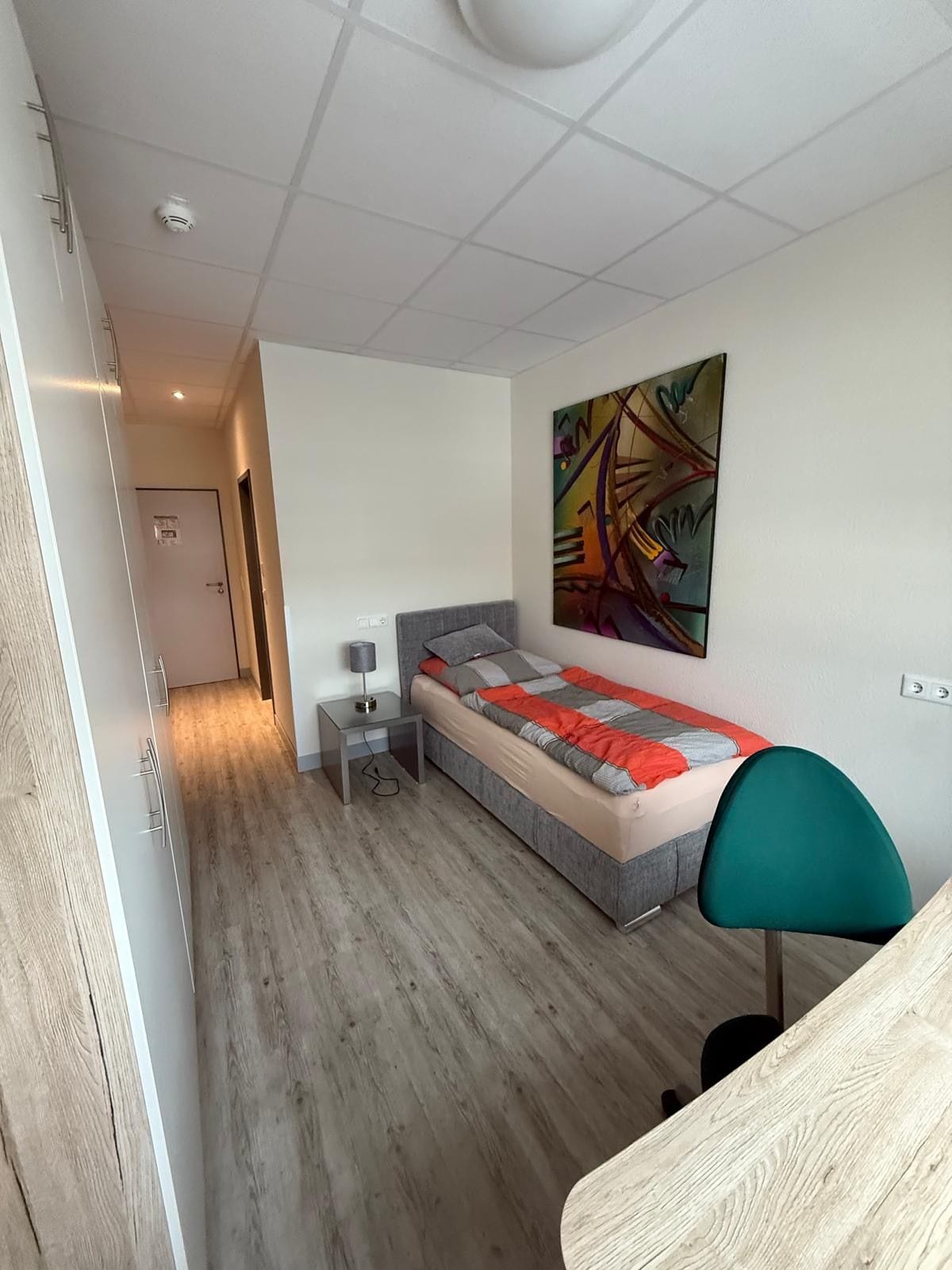 A modern bedroom with a bed, bedside table, lamp, and a colorful abstract painting on the wall.