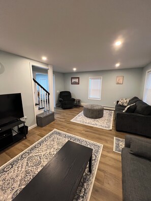 A living room with a TV, sofa, ottoman, and a staircase leading to another room. - “Twin Haven Manistique 2BR Retreat” (Manistique)