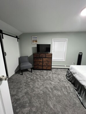 Ein Schlafzimmer mit grauem Teppich, einem grauen Sessel, einem Holzschrank, einem Fernseher darauf, einem Fenster mit Jalousien und einem Bett mit gemustertem Überwurf.