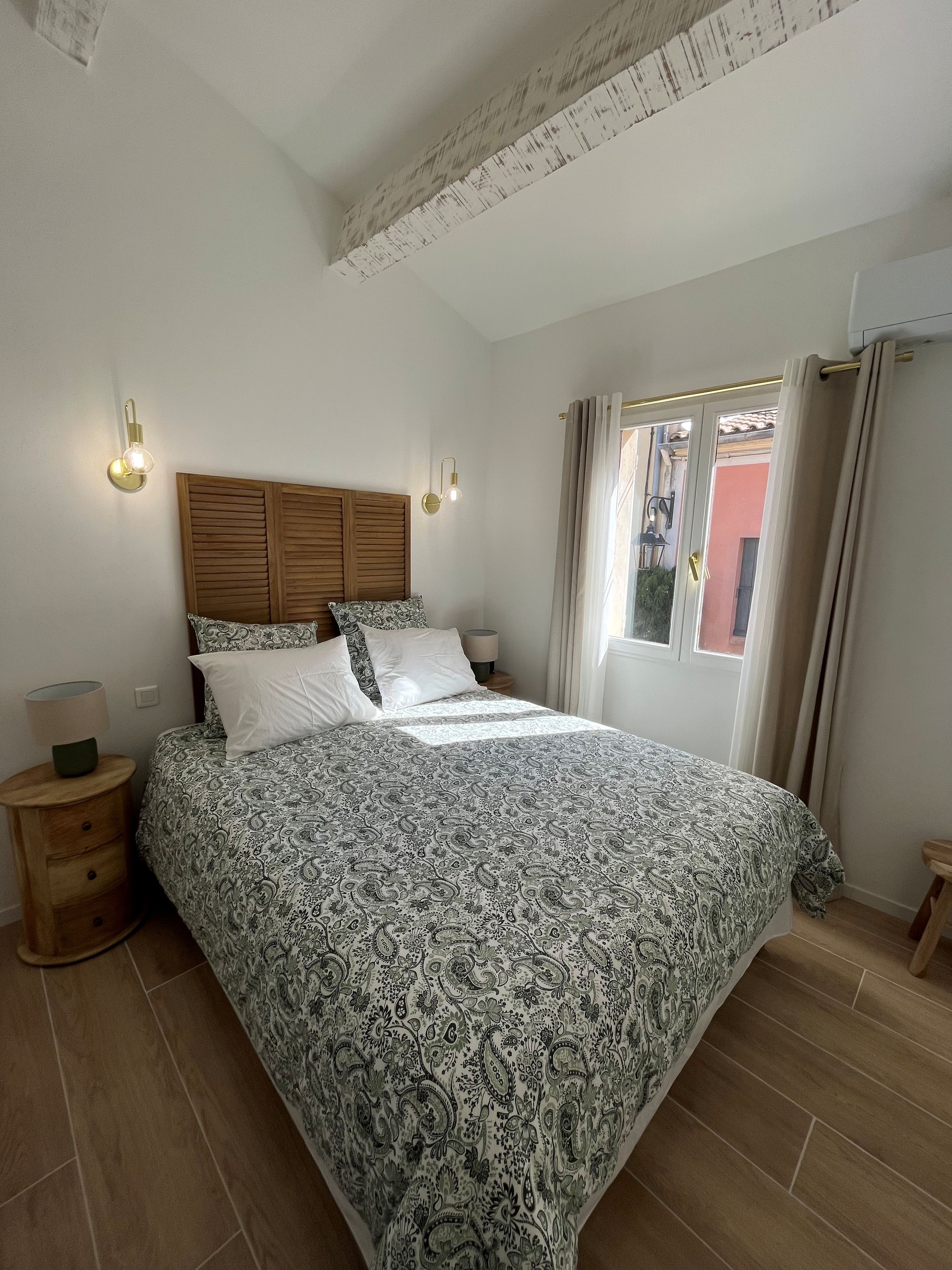 Een slaapkamer met een houten hoofdbord, een bed met een decoratieve spreidek, twee bedlampjes, een nachtkastje en een raam met gordijnen.