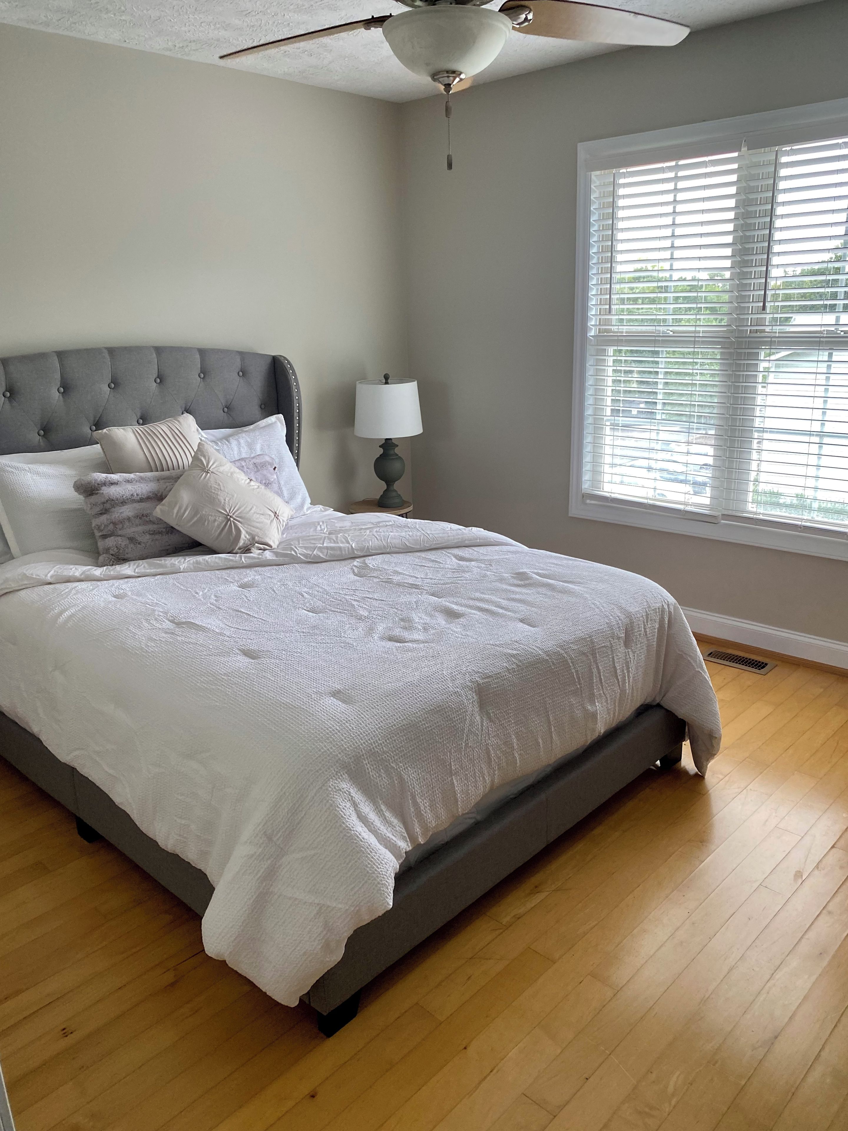 A bedroom with a bed, a ceiling fan, a bedside table with a lamp, and a window with blinds.