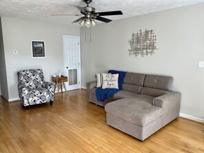 Ein Wohnzimmer mit einem Ecksofa, einem gemusterten Sessel, einem kleinen Holztisch, einem Deckenventilator und einem Wandkunstwerk.