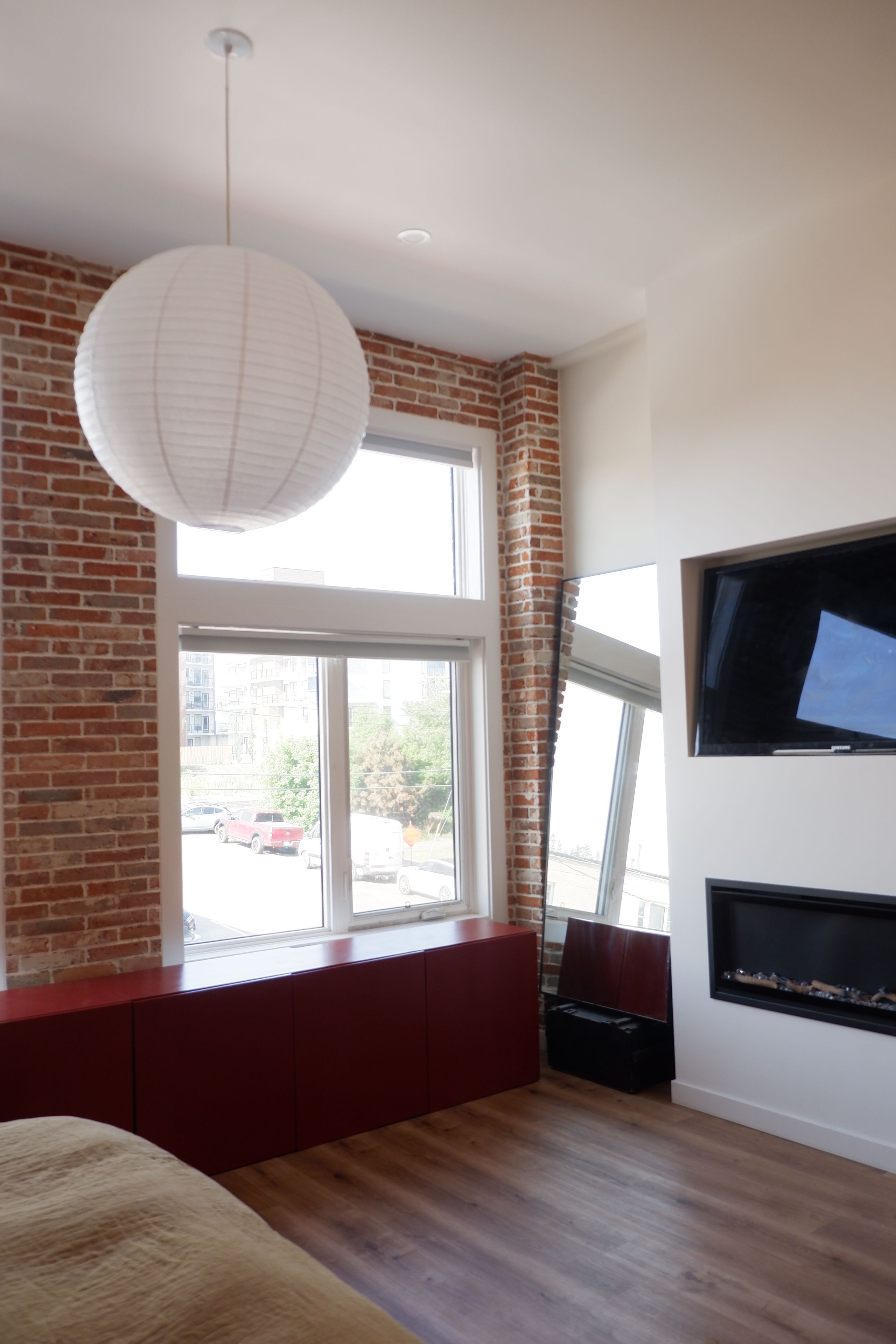 A modern room with a brick wall, a large window, a red bench, a flat-screen TV mounted on the wall, and a hanging light fixture.