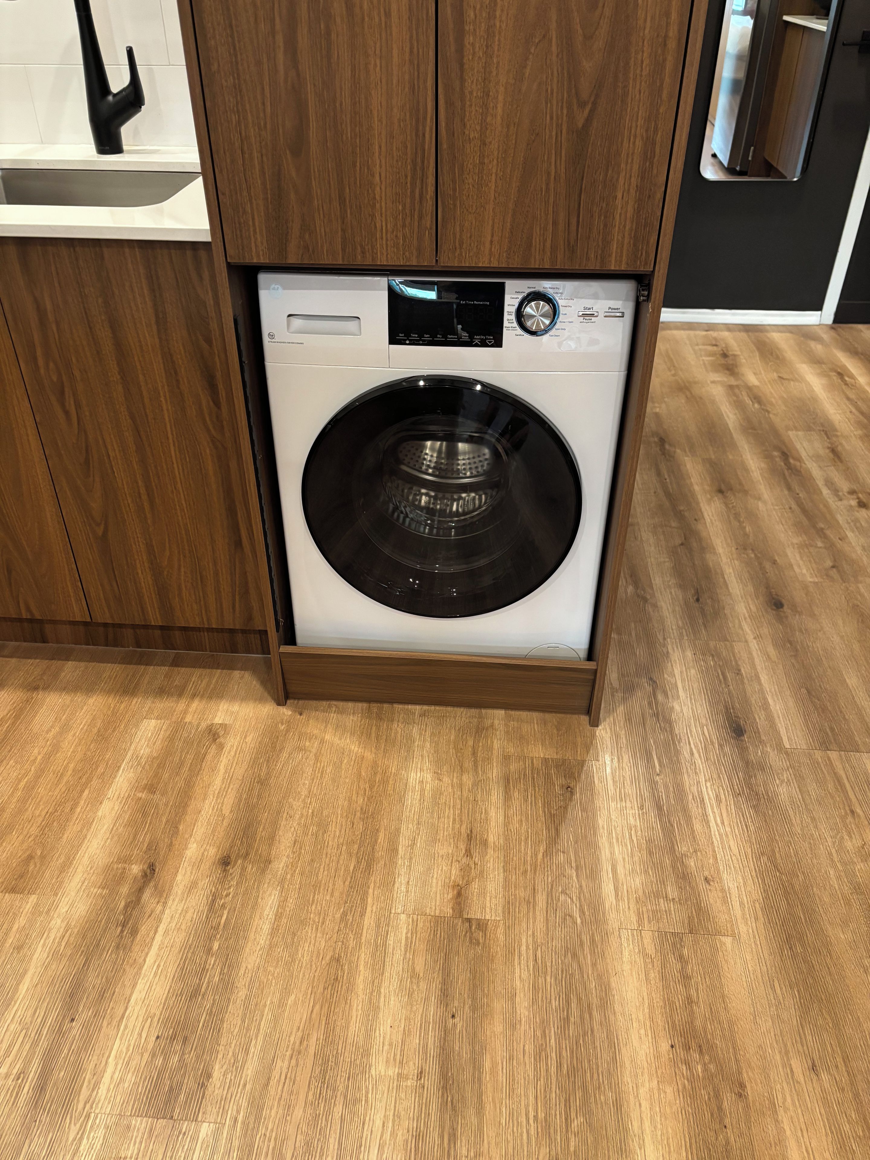 A front-loading washing machine in a kitchen with wooden cabinets and a wooden floor.
