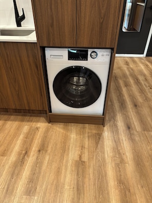 A front-loading washing machine in a kitchen with wooden cabinets and a wooden floor.
